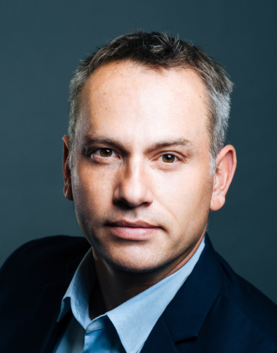 A man with short gray hair wearing a dark suit jacket and light blue shirt against a gray background.